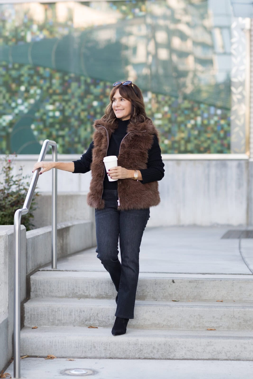 Susie wearing fall outfit with brown faux fur vest, black sweater, grey jeans and black boots holding a coffee cup.