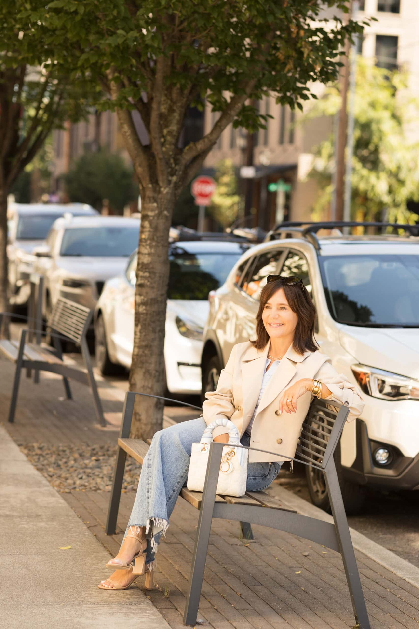 Susie sitting on bench wearing light was denim with cropped trench coat and white lady dior bag with scarf on handle