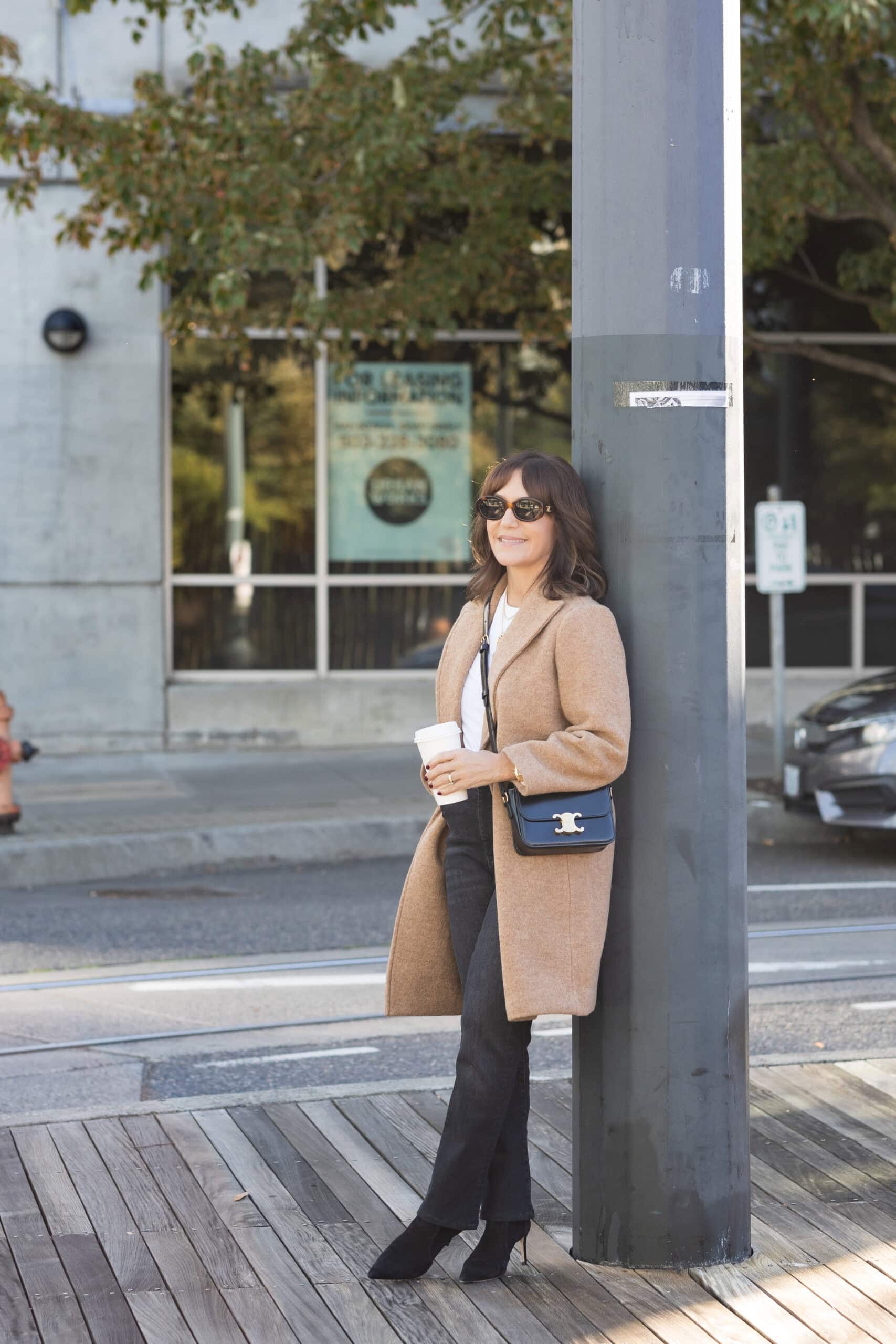 So Susie outdoors in a chic fall outfit with camel coat, layered sweater, and wide-leg jeans.