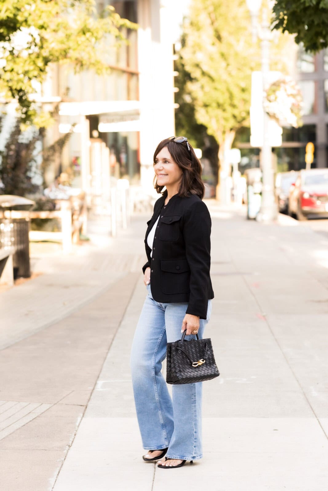 Susie in light wash jeans, black strappy heels, black jacket, with black leather woven handbag.