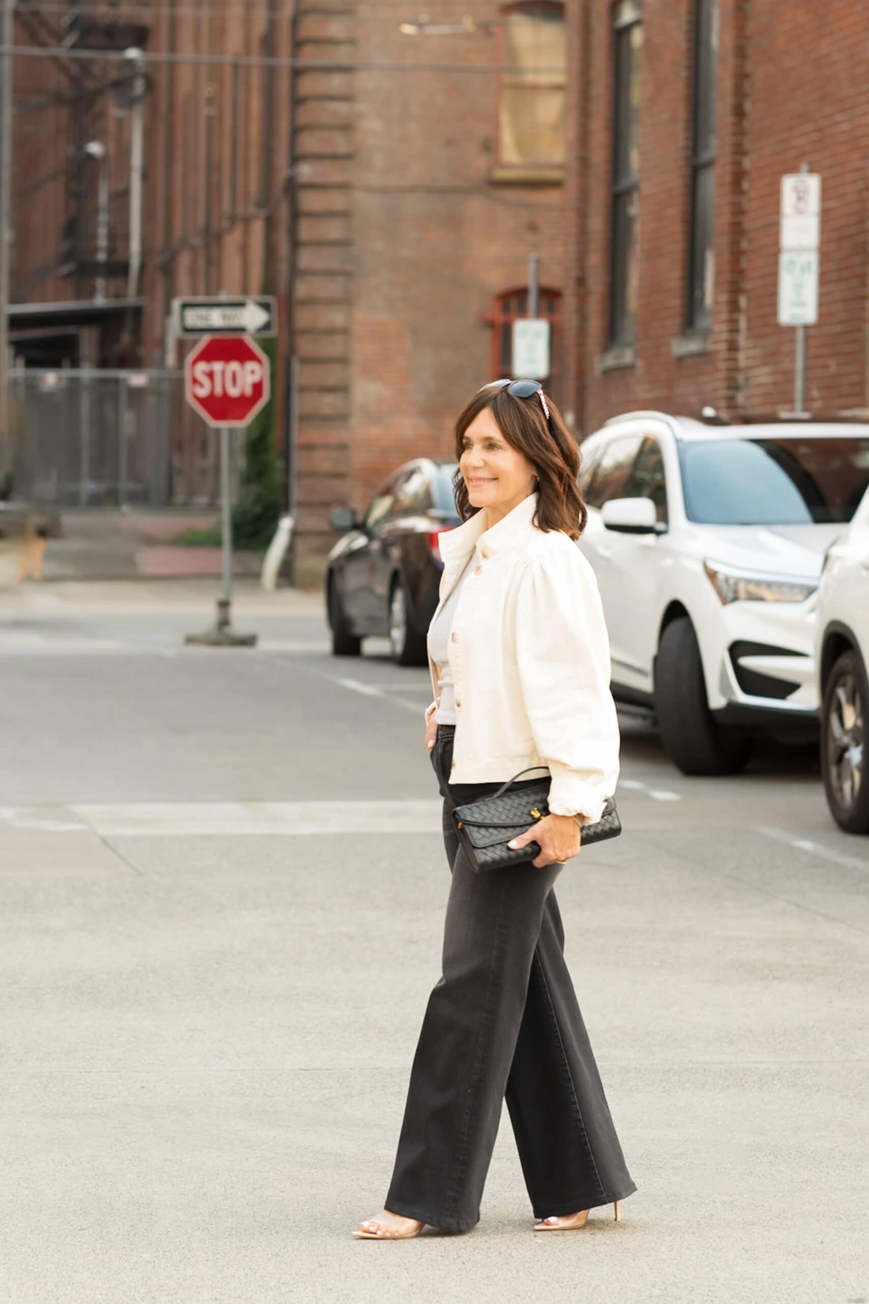 Susie in a white jacket with black wide leg jeans and a black leather woven clutch.