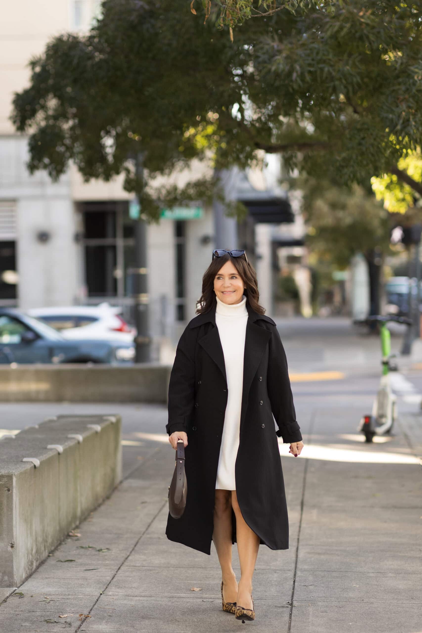 Susie walking wearing black trench with ivory sweater dress and jimmy choo shoes and purse